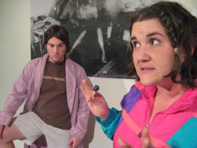 A woman in a brightly colored tracksuit is eating a sucker and talking to a man in a bathrobe. On the wall behind them is a photo of a spontaneous human combustion.