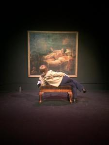 A female art museum security guard reclines on an upholstered bench in front of a Rembrandt’s painting of Danae. The guard reclines in the same manner as Danae in the painting.