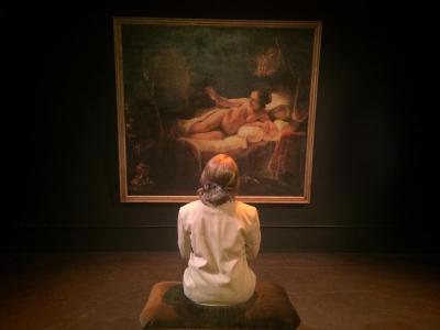 A female art museum security guard sits on an upholstered bench facing Rembrandt’s painting of Danae.