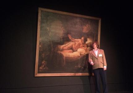 A female art museum security guard stands next to Rembrandt’s painting of Danae. She leans in close and matches the eye line of the painted woman in an attempt to determine what she might be looking at.