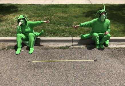 Two people wearing bright green grasshopper costumes are sitting on a curb. On the ground in front of them is a tape measure showing that they are six feet apart. They are reaching a had towards each other. Both of them have a light green handkerchief covering their mouth and nose.