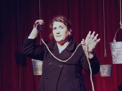 A woman in a peacoat and fingerless gloves holds rope tied to a spoon in a threatening manner. Several metal buckets hang from above.