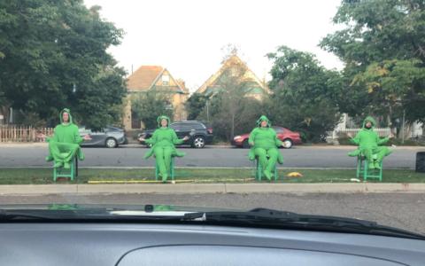 A picture from inside of a car. At the bottom of the picture is the dashboard. In the distance, four people dressed in bright green grasshopper costumes are sitting in green chairs with their hands as though they are meditating. Behind them is a street with cars, houses, and trees.