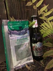 A plastic package of very moldy and green provolone cheese, next to a bottle of beer and a branch of green leaves. 