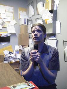 A man sits at a desk singing earnestly into a microphone. The walls behind him are covered in papers, letters, and postcards.