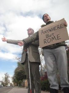 A disheveled man dressed for winter is holding a cardboard sign that reads "anywhere but rome" his other hand is thumb out hitchhiking. To his left there is a man in a suit with dark glasses holding a white cane, also with his thumb up, hitchhiking. 