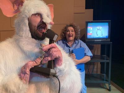 In the foreground a Giant white Rat is yelling into a microphone.  Behind him, a scientist wearing a blue jumper and holding a blue clipboard, is smiling. Next to her a television on a cart is showing a ring that is being sold on the home shopping network.