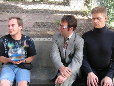 Three awkward men sit in front of a chainlink fence. On the left is a smiling man with a space t-shirt on he is holding a fish bowl with a goldfish. In the center, a man in a suit and large glasses purses his lips. On the right is an intense man with a buzz cut and black turtleneck. 