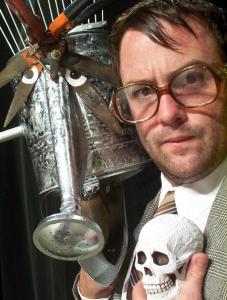 Close up of a  man with large glasses holding a small skull. Next to him is a puppet made from a watering can with eyes and rake hair.
