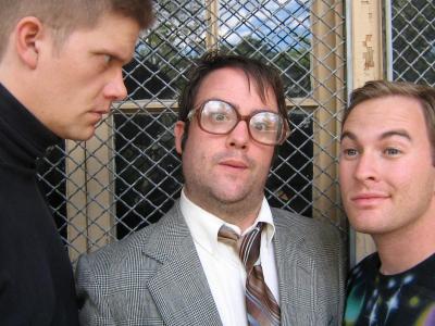 Close-up of three awkward men in front of a window grate. On the left is an intense man with a buzz cut and black turtleneck. In the center, a man in a suit and large glasses purses his lips. On the right is a smiling man with a space t-shirt on