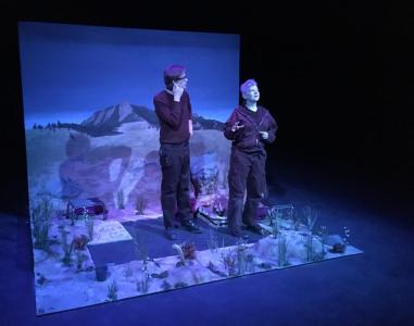 Two people wearing black stand in a partially finished scene of a prairie with distant mountains. Some of the flooring is bare. One person talks out as the other looks at them. Everything is bathed in blue light.