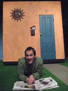 A man smiles while lying belly down on astroturf, an open newspaper in front of him. In the background is a small stuccoed house with a blue door and a full mailbox.