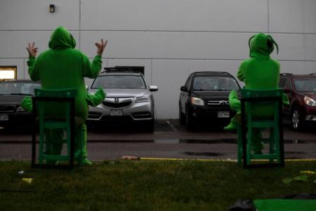 In the foreground, on a strip of grass, two people in bright green grasshopper costumes are sitting in green chairs with their backs to the camera. They are facing a row of cars parked in a parking lot in front of a white building. It is rainy.