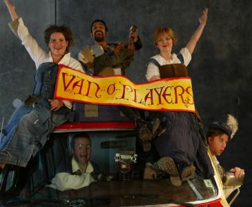 Three people sit on top of a van. Two are holding a banner that says “van-o-players” while the third happily holds a stringed instrument. Inside the van, through the windshield, is a crying man. Popping his head out of a side window is a surprised-looking man in a feathered cap.