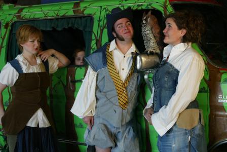 A van painted like a forest provides the backdrop. Standing on either side of the sideview mirror are two flirting people. There is a plastic owl on the mirror. To the side is a woman glaring at the couple. In the corner of the window, part of a head peaks out. Everyone is wearing makeshift Shakespearean clothing.