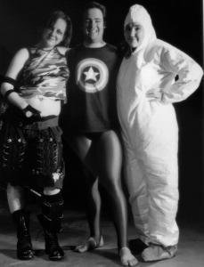 Three friends, one dressed in a hazmat suit, one dressed in apocalyptic attire, and one dressed as a superhero and wearing a Captain America t-shirt pose and smile out towards the camera. 