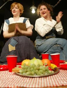 In the foreground, a platter of fruit and some red plastic cups sit on a red checkered tablecloth. Behind, two women in makeshift Shakespearean clothes smile and clap. There is bare bulb hanging between them.