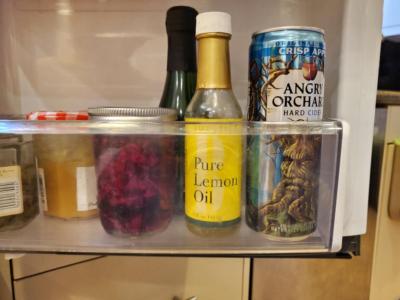 Picture of the inside of a refrigerator door. Several items are lines up, including a bottle of Pure Lemon Oil and a can of Angry Orchard Hard Cider.