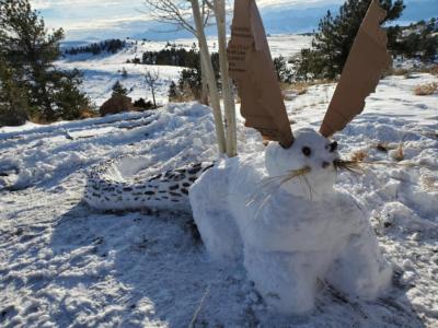 Snow Rabigator-A snow creature sits on a snowy hill with trees and mountains behind. It looks somewhat like a rabbit, with straw whiskers and large cardboard ears, but has a long snake-like tail behind it, decorated with bits of bark.