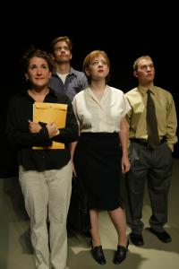 Four people wearing business casual attire stand together. All of them are looking up. One holds a briefcase, another holds a large yellow envelope.