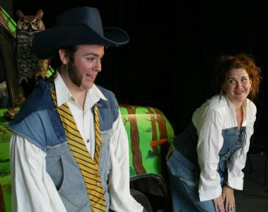A man with sideburns and a large cowboy hat is talking. Over his shoulder is a plastic owl staring at the camera. A woman standing with her hands on her knees is smiling at the man.