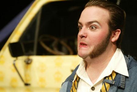 Close-up of a man with large sideburns. He is bug-eyed and grimacing. In the background, out of focus, in the front of a yellow van.