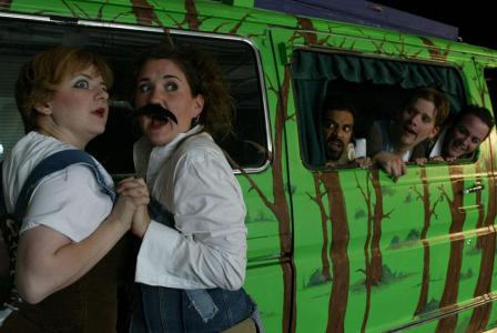 Two women clasp hands as they look out, wide-eyed. One of the women wears a fake mustache. Behind them, peaking out of the window of a van that has been painted like a forest, three men are watching.