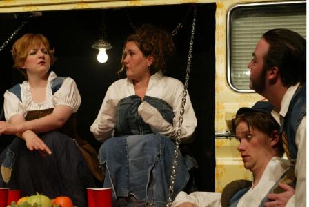 Two women sit in a panel that has been flipped down in the side of a yellow van. There are chains holding the panel in place. One woman looks confused. The other has her hands covered by her sleeves. On the side of the van, two men are sitting in profile.