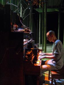 A man plays a piano that is covered with tape recorders. Behind him a forest is illuminated and green.