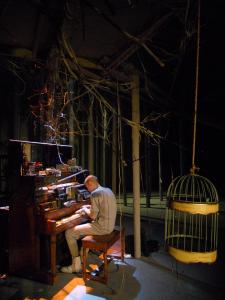 An empty birdcage hangs in the foreground. Behind it a man plays a piano covered in tape recorders.