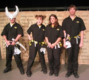 Four confused people wearing utility suits and utility belts with origami paper props hanging off of them are standing in a line. One of the people is wearing a huge Viking horned helmet made of paper. The helmet is covered in the word 'helmet.'