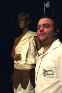 A man wearing black earphones and a white polo with a large logo on the pocket stands in front of a mannequin in a white toga 