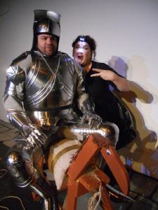 A man dressed in a suit of armor sits on a wooden horse. Behind him a woman with a head lamp photo bombs the picture.