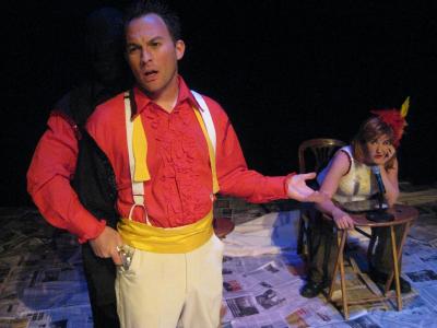 A man in a brightly colored suit stands talking. One of his arms appears to be separated from his body and is putting a toy gun in his pocket. Seated in the background, a woman dressed as a magician’s assistant but wearing mechanics pants looks unimpressed.