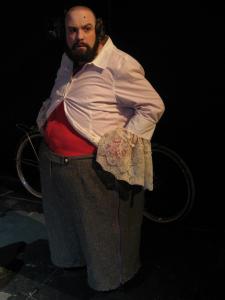 A large bald man with a big beard and a beauty mark on his forehead looks out suspiciously. He wears grey trousers and a white shirt with long lace cuffs. The shirt is open, revealing red long underwear over his belly.