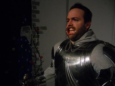 A man dressed in a suite of armor stands in front of a painting of a wizard. The man is wearing the lip expanders you might wear at the dentist’s office during a procedure.