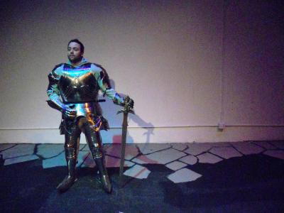 A man stands in front of a white wall dressed in a suit of armor. He holds a large sword by his side.