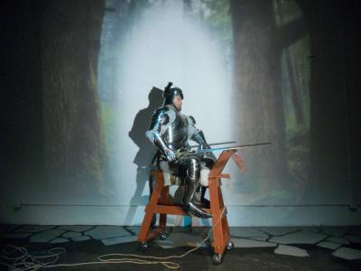 A man sits on a wooden horse in a suit of armor carrying a large sword. A forest is projected on the wall behind him.