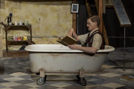 A mustached man sits in a claw-footed bathtub on casters. He is wearing a short-sleeved shirt and a sweater vest and he is holding an open book in front of him. The bathtub is center stage and sits on a grey and white checkerboard floor. In the background there is a wooden cart with tea cups and boxes of tea on it.
