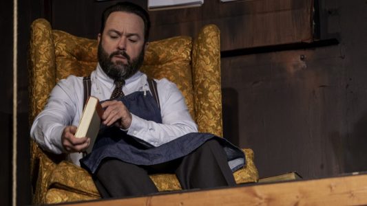 A bearded man in a button-up shirt, tie, and denim apron looks at the spine of a book as he sits in a orange floral wingback chair.