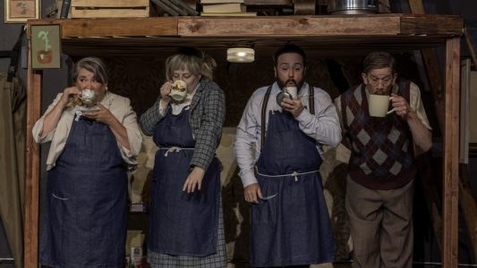 Four people are standing, jammed in under a wooden platform. They each are drinking out of different teacups. Three are standing up straight and wearing denim aprons over their business clothes. The fourth, much taller, has to bend down and is wearing slacks and a sweater vest. Above them on the platform are piles of books. There are a few bad paintings of a plant hung around them.