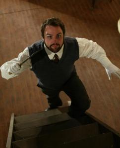 A view down a staircase of a man with dark hair and a beard holding a knife in his left hand raised in front of him menacingly.