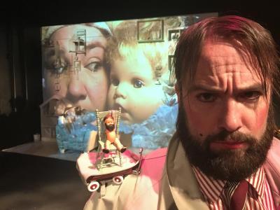 Close-up of an unhappy man with a beard and a beauty mark. On his shoulder there is a tiny doll with a beard sitting on a tiny chair sitting in a tiny red wagon. In the background, projected on the wall, is an extreme close-up of a person looking at a baby doll head.