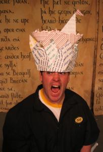 A screaming person is wearing a crown made of paper that has the word water printed all over it. On top of that crown is an origami boat made of paper that has the word boat printed all over it.