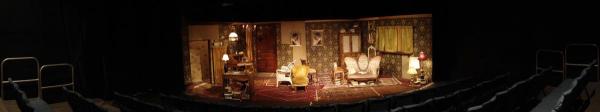 A panoramic shot from the back of the room shows the whole stage. The set has a patchwork quality to it, mixing different carpets, fabrics, and wallpapers. It looks cluttered.