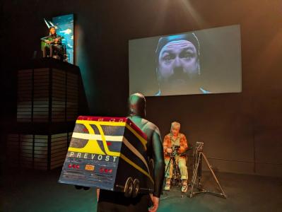 In the foreground there is a person wearing a colorful bus built around their waist. Their face is being projected onto a screen that is on the wall in front of them. Below that screen sits a woman surrounded by 3 video cameras pointed in different directions. To the left of these too we see a person sitting on top of a tall box structure, holding a green guitar, representing the Sears Tower.