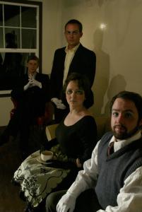 A strange portrait of four emotionless people. They all wear gloves and dark clothes. They have a lot of eye make-up, giving a sunken eye effect. In the background is a window.