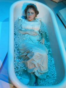 Mary Shelley, laid back in a bathtub full of ice cubes, wearing only her corset and shift, looks seriously into the camera.