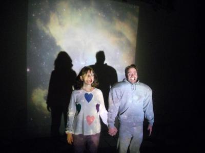 Man and woman stand in front of a projected galaxy, both are wearing the lip expanders you might wear at the dentist’s office during a procedure. The man is in a gray sweatsuit with shoulder pads and the woman is wearing a white shirt with colorful hearts.