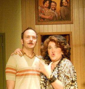 A woman with large bouffant hair and a leopard-print caftan talks, with are arm around the shoulders of a mustachioed man. Behind them is wood paneling and a family portrait featuring the woman.